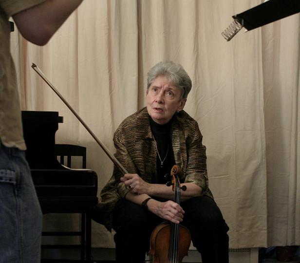 Sally Thomas is sitting with a violin and bow in hand, looking up at a student standing outside of frame. Thomas' facial expression is one of attention and perhaps inquiry or discussion. The room has a simple and functional decor, with a plain curtain backdrop and a music stand, which suggests a focus on music practice.